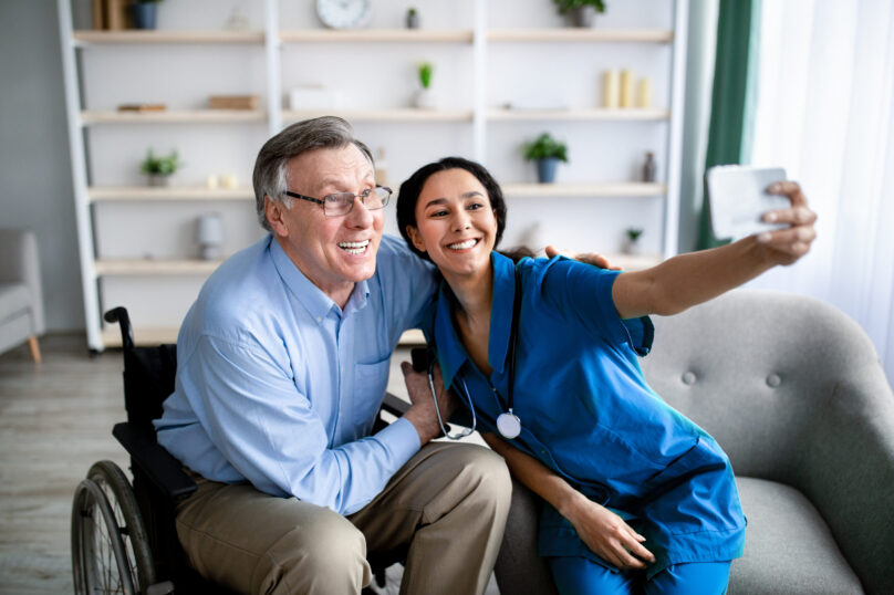 Cheerful impaired senior man in wheelchair taking selfie with young female doctor at retirement home Cheerful impaired senior man in wheelchair taking selfie with young female doctor at retirement home. Disabled older patient making photo with nurse, having fun together indoors
