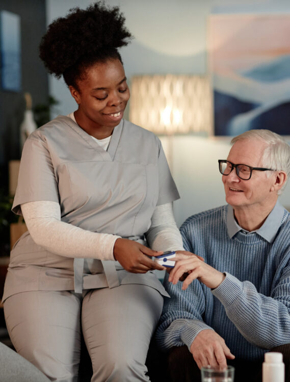 Supportive Black female social worker visiting upbeat elderly man checking up oxygen saturation in blood with pulse oximeter, while sitting together on couch providing medical assistance at home